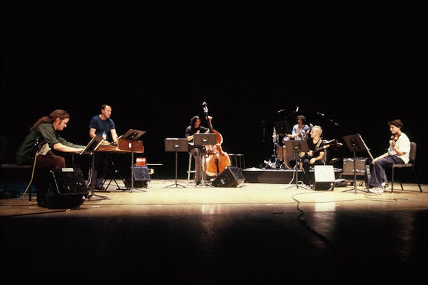 Several performers sit on chairs arranged in a semi-circle within a dark stage setting. The performers play instruments such as electric guitar, bass, drums, xylophone, and violin. 