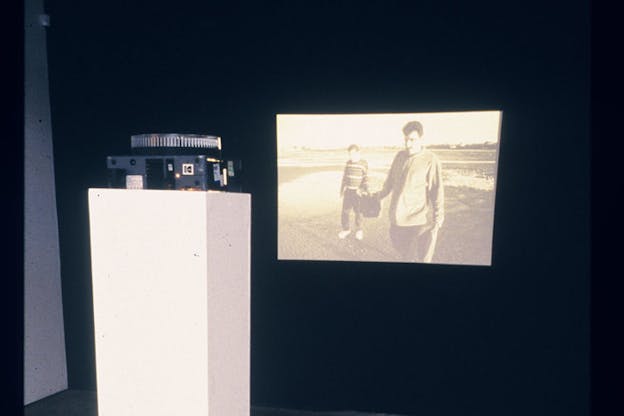 A projector on a white square pedestal faces a sepia toned image of two people standing on a wetland. The rest of the wall is black. 