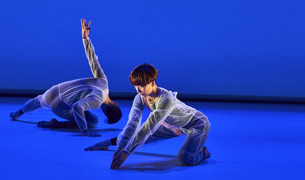 Two performers dance on a stage awash in deep blue light, illuminted warmly from the sides. Hsiao-Jou Tang is centered in the frame with her body facing towards the left, kneeling on one knee with the other leg extended straight out to her side. Her torso is pitched forward and her arms are extended before her, palms touching as her fingertips touch the floor. Further back, to Tang's right, Alex Springer is posed also on one knee with his body and head facing away from the camera. His torso is leaned far to the right with his head almost touching the floor. Both are dressed in sheer white jumpsuits over black underclothing.