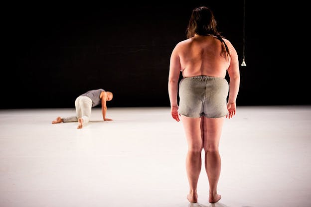 In the front mid-right of the stage, a performer wearing nothing but shorts stands with their back turned while downstage to the left, an out-of-focus performer crawls on the floor.