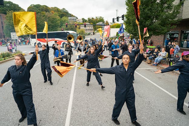 People dressed in black overalls holding colorful flags and musical instruments parade in lines of three.