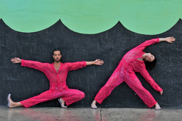 Two performers in hot pink lace jumpsuits and jelly sandals strike poses against a gray wall painted with mint green scallop print.