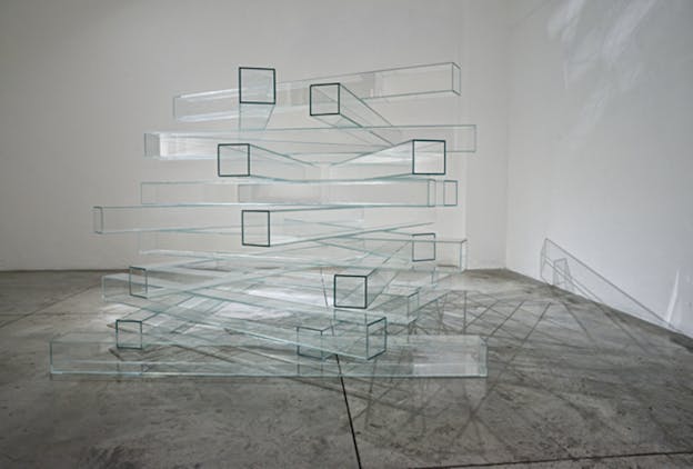 Dozens of clear glass square tubes are criss-crossed on top of one another like a tower. Light entering from above creates patterns of shadow on the floor and wall. The installation sits on a concrete floor against an off-white background. 