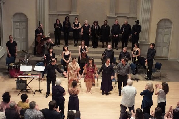 Performers, ensemble, and choir bow before an applauding audience, the front row standing in ovation.