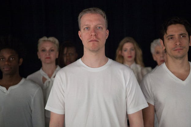 Close-up of persons standing shoulder-to-shoulder in three rows all wearing white shirts and facing forwards, the person in the front center's face is focused and the others' are blurred. 