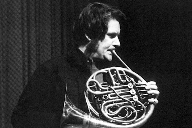 A close up black and white image of Gordon Mumma playing a horn in front of a curtained space. He wears a dark shirt and looks intently into the distance. 