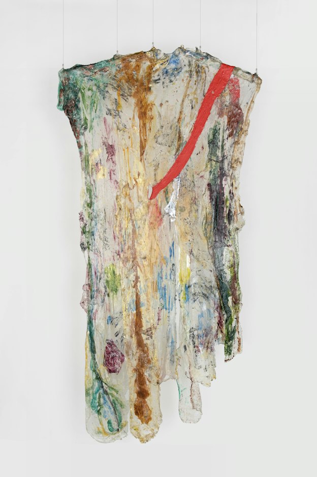 A hanging sculpture made of transparent plastic draped over two, short wooden rods, with painted areas. The rods are tilted slightly, resembling the arms of a clothes hanger. The plastic ends are cut in rounded forms. Dark green, red, brown, and magenta paint areas are visible on the plastic. A strip of red plastic starts at the top right and drapes across the face of the transparent piece, the bottom end coming through a hole in the center of the piece towards the back. 