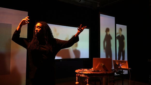 A photograph of Pamela Z performing, standing with her arms upraised and her eyes closed. Behind her, wall projections show three people clad in black gesturing and interacting with one another. While Pamela Z is illuminated with orange light, the rest of the scene (including a table with laptops on it) is shrouded in darkness. 