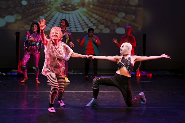 Two performers in front center-of-stage clasp hands, one reaches outwards and one in a head mask kneels and reaches out to the side. Behind them, four performers dance in front of a projection of gold dots.