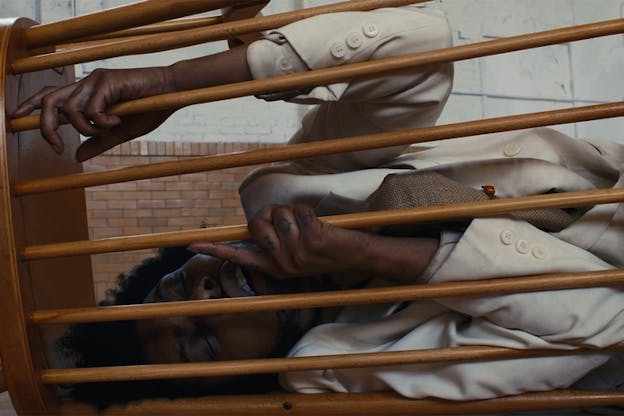 A close up of a performer and a chair laying horizontally on the ground. The performer stands with their eyes closed behind the chair's upper part which consits of thin parallel wooden connectors.