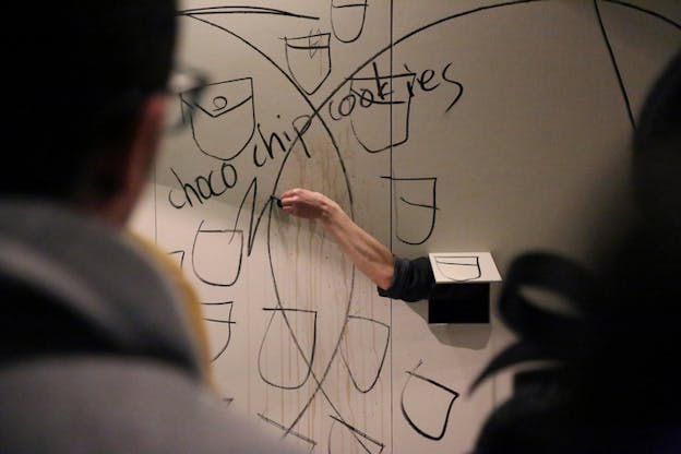 A hand holding a black marker reaches out from a cut square on a wall as it doodles underneath the written words 