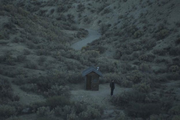 A video still of a person at dawn, standing outside a small structure, facing away from the structure and looking towards the distance. The structure is wood paneled and has a triangle roof. It is only slightly taller than the figure. All around this, numerous small scrubs are scattered across a sandy landscape. In the distant background, a winding pathway is visible. The entire image is hued with a cool colors and there is minimal bright light.  