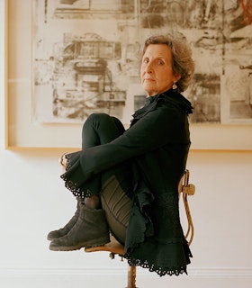 A portrait of Trisha Brown sitting sideways on a chair with her knees pulled up towards her chest. She turns to the camera directly. She has short grey hair and wears a black shirt with patterns cut out by the sleeves and bottom edge, sage green pants, and light brown boots. Behind her, there is a white wall and the bottom left edge of a framed light green abstract image.