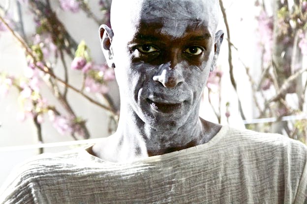 In a performance, David Thomson stares at the camera with his face and neck painted chalky white, wearing a white textured top. Behind him, there are pink flowered branches against a white background.