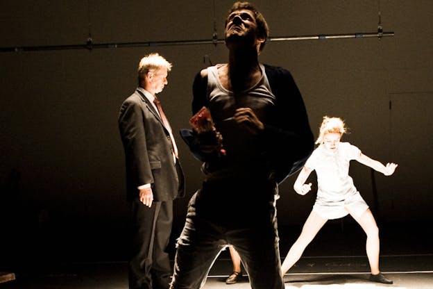 A performance still of three performers in a bare, darkened space. In the foreground, a man dressed in pants and a white tank top and holding a colored plastic bag squeezes their hangs into fists and turns their head upwards with their eyes squeezed shut. Behind this performer, another performer, dressed all in white and illuminated brightly, lunges sideways and looks downwards while another performer in a suit looks intently at them. 