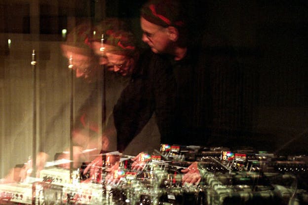 A blurred image of Knittel in front of musical equipment. He wears a dark shirt and a hat with a red stripe. The image of him leaning over the musical equipment is blurred so that he appears three times.