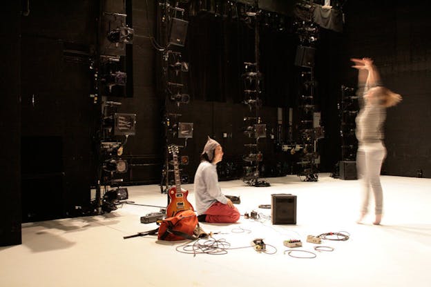 Person sits on a brightly lit stage, a guitar behind them, watching a dancer relevé, their body blurred in motion. 