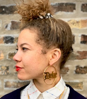 A profile portrait of Anna Martine Whitehead in a white collared shirt. They are wearing red lipstick and a hoop earring with the word 'dyke' spelled in the middle.