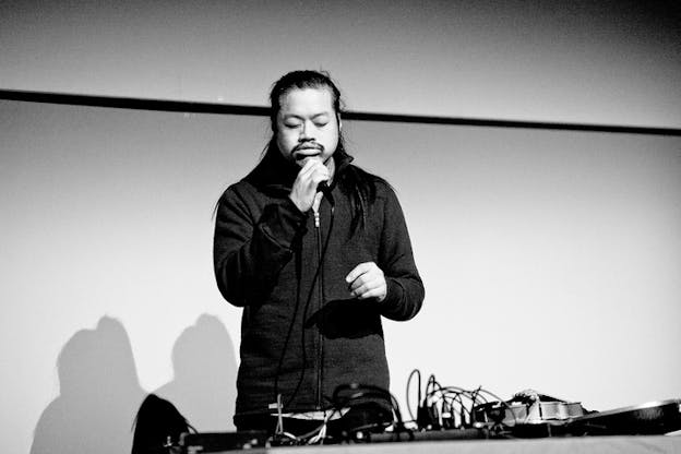 Black and white photograph of Spencer Yeh singing on a microphone behind a desk supporting a pedal board with various cables.
