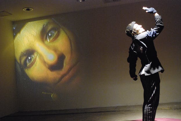 Mac Low performs, standing with her hand raised above her head, looking towards it. She wears all black and has black straps across her head. On the wall behind her, there is a orange tinted projection of a close up of her face. 