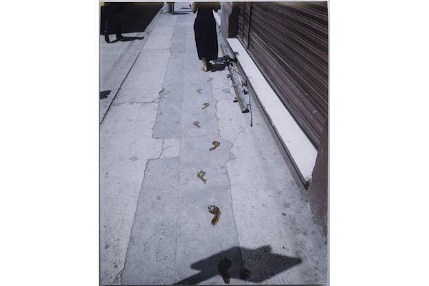 A barefoot figure clad in black leaves behind on the street red footprints as they walk. 