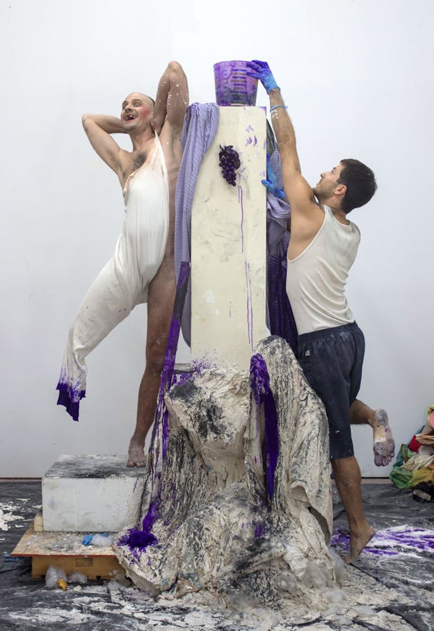 Two performers stand around a white rectangle column. On top of the column there is a clear plastic bucket with purple paint in it and below that, a bunch of purple painted grapes. The paint is visible dripping in various places throughout the installation, including on the bottom of the column which is covered in draped white and gray plaster. One performer wears thin white fabric which covers most of their torso and one of their legs. The bottom of this fabric drips with purple paint. This performer stands on one leg and holds the back of their next with their hands. They have an emphatic expression, pink makeup of their cheeks, and are missing a few teeth. Matt Savitsky wears a white tank top,  blue shorts, and blue plastic gloves. He reaches with one gloved hand toward to the bucket of paint. 