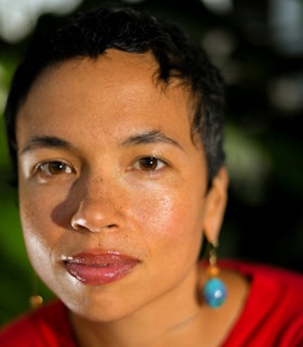 Holland Andrews looks directly into the camera. Their face is in sharp focus against a softly blurred background. They are wearing a bright red top and bright blue pendular earrings.