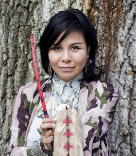 A portrait of Laura Ortman against a tree trunk. She wears a light blue patterned shirt and a pink, purple, and green patterned cardigan, red heart earrings, and light blue and black eyeliner. She holds a red and black patterned wand and a tan and red patterned cylinder.  