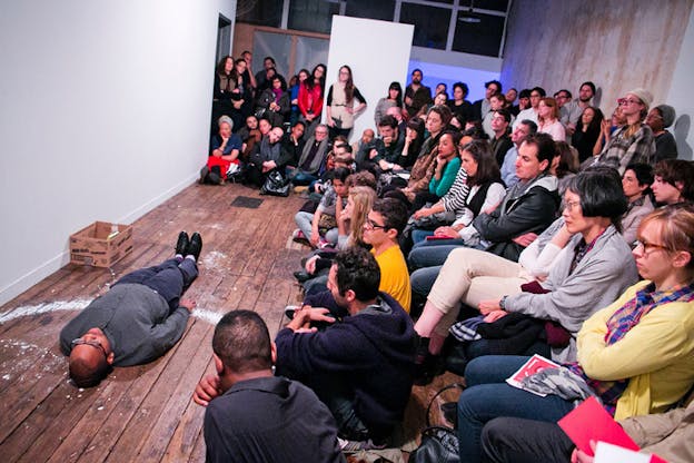 A performer lays with their back on the wooden floor near a white wall surrounded by an audience watching them.