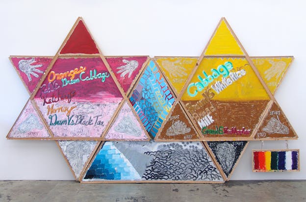 Two star-shaped freestanding panels connected by a diamond-shaped panel. The left panel is painted in shades of pink and has text that reads: “Oranges, Red vs Green Cabbage, Ketchup, Honey, Green vs Black Tea.” The right panel is painted in shades of yellow and brown and has text that reads: “Cabbage, Brown vs White Rice, Bread, Milk, Green vs Red Apples.” Other wooden panels adjoin the two stars and are painted with rhomboids, squiggles, and colored bars.