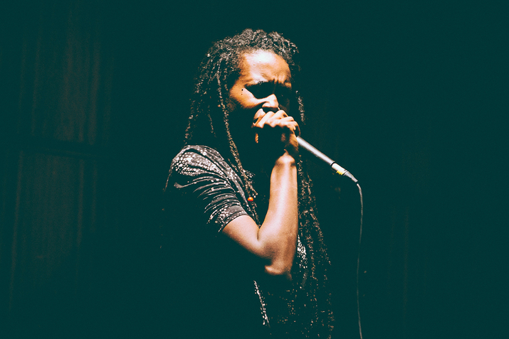 A performer most of their characteristics shadowed holds a microphone stand close to them singing. They wear a black with white splattered dots top and long dreadlocks.