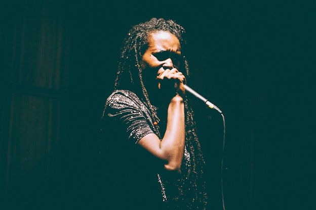 A performer most of their characteristics shadowed holds a microphone stand close to them singing. They wear a black with white splattered dots top and long dreadlocks.