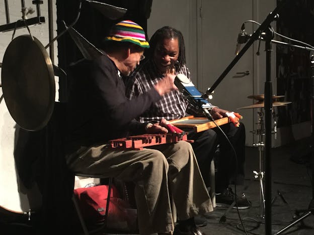 Newman Taylor Baker sits onstage next to Warren Smith, surrounded by microphones and cymbals. They are smiling at each other and holding their instruments in their laps.