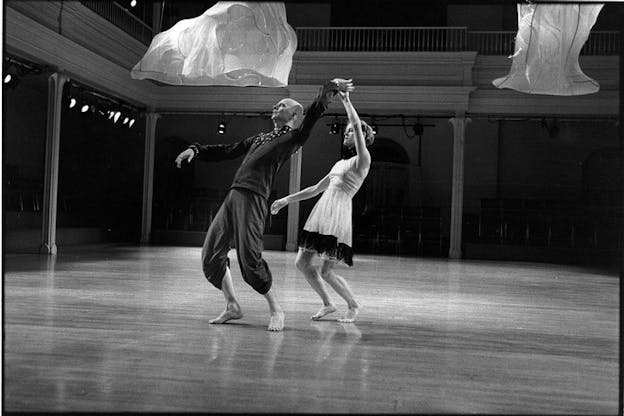 Black and white image of two performers the one behind the other hold hands turned upwards. 