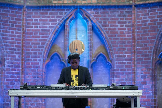 A DJ dressed in a yellow shirt and black dress jacket stands behind a DJ controller en face to the viewer. Behind the set up is located a brick wall with an almond shaped center of blue and gold wall centering the musician.