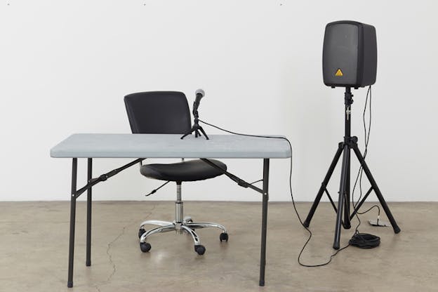 A gray desk hosts a microphone on it.Behind it a black chair and on the left side a black acoustic system on a tripod. Everything is connected through cables.