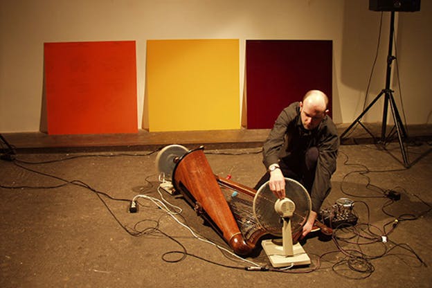 Davies positioning a fan beside a wooden harp placed on the floor in a warmly-lit room with three squares of orange, yellow, and maroon positioned against the back wall.