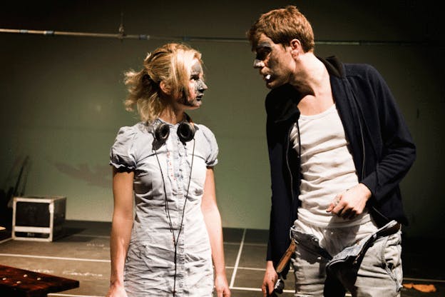 Two performers stare intently at each other with their faces covered in mud. One performer wears a white dress and headphones and the other has a tank top, sweatshirt, and unzipped pants. 