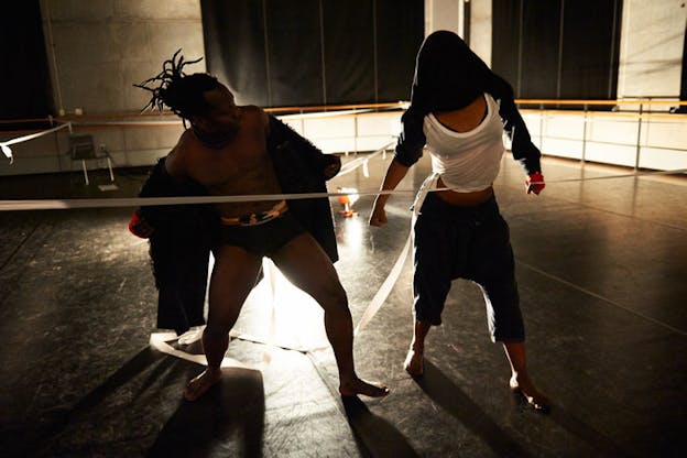 Two performers stand behind a white line across their midriffs. The one on the left is dressed in black underwear and an open black robe looking towards the person on the right dressed in dark pants, a white tank top and a black top which covers their face. 