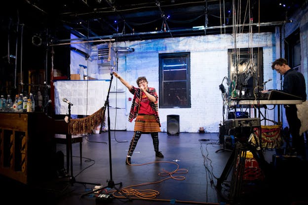 Performer sings into a microphone and raises their right arm on a chord-cluttered stage in front of a white-painted brick wall faintly lit water blue. 