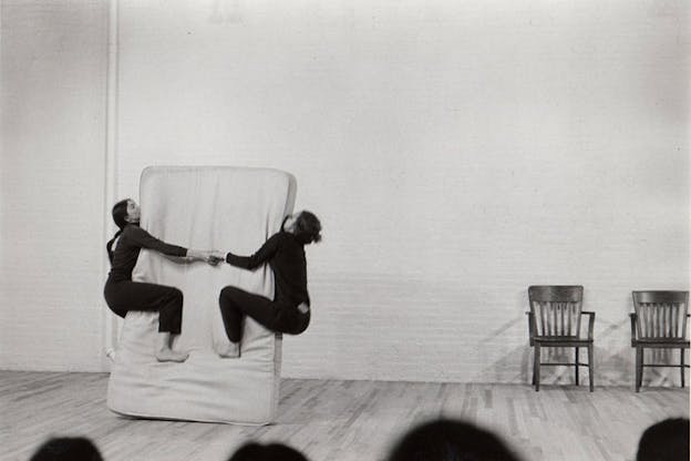 Black and white still of two performers in matching black turtlenecks and pants hanging onto the sides of an upright, tilted mattress, their hands interlocked.