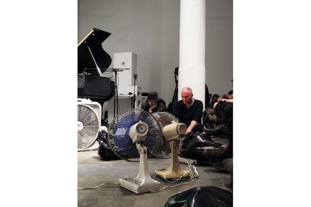 People sitted on the floor surround three fans. In the back left behind a fan the side of a piano can be viewed.