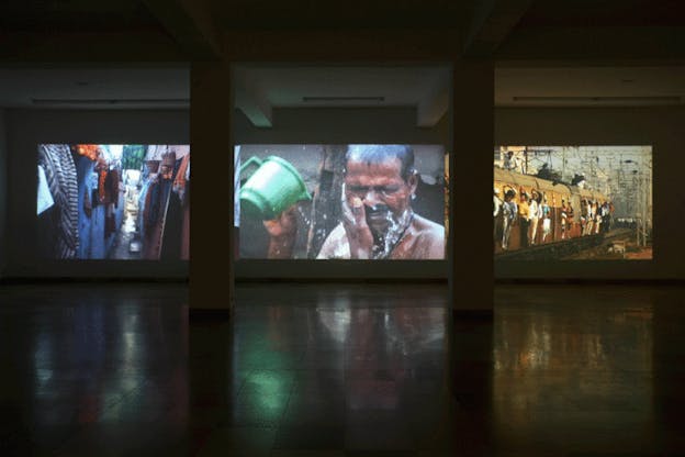 An installation image of a large screen broken into thirds in a sparse white room. Two white columns are situated perfectly in front of the lines where the screen has been segmented. On the left of the screen, there is the image of a thin street, in the middle there is the image of a man washing his face with water from a green bucket, and on the right, there is an image of passengers leaning out of an orange train.  