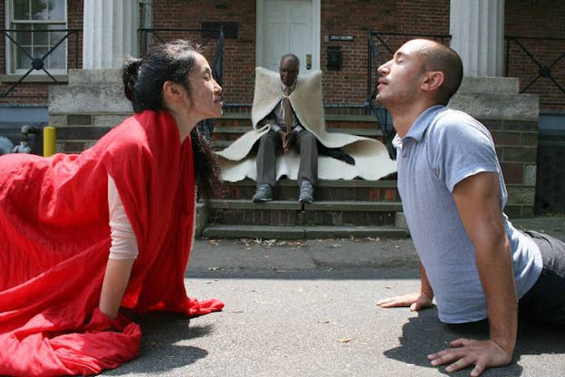 Two performers rest on their hands and knees on the sidwalk. The performer on the left wears a red draped fabric. The performer on the right wears a grey shirt and black pants. Both performers close their eyes and angle their heads towards each other. A third performer wears a draped cream cloth and sits on a staricase behind them. The staircase leads to a brick building with white columns.
