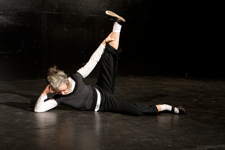 Performer leans on a black stage floor with their head propped against one elbow, holding one leg outstretched in a ninety-degree angle.