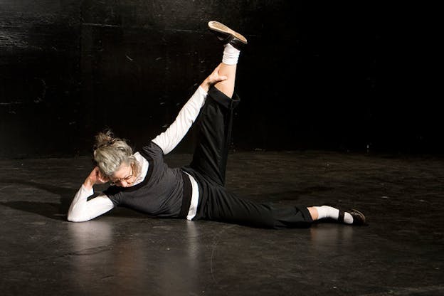 Performer leans on a black stage floor with their head propped against one elbow, holding one leg outstretched in a ninety-degree angle.