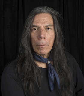 Portrait of Brad Kahlhamer dressed in a black shirt, a small blue scarf around the neck and straight long graying hair.