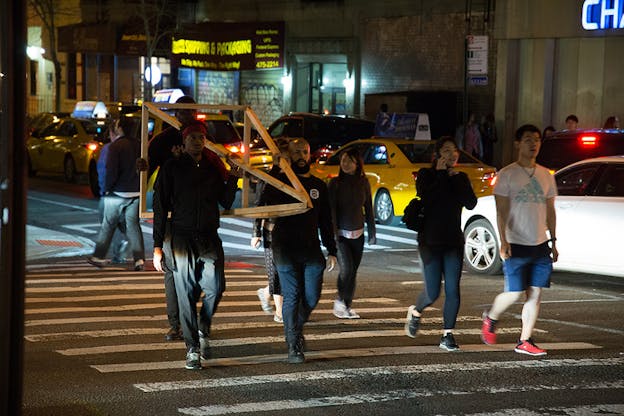 Performers walk across a New York City crosswalk at night. Three performers hold a structure of wooden beams between them. All performers are looking forwards. 