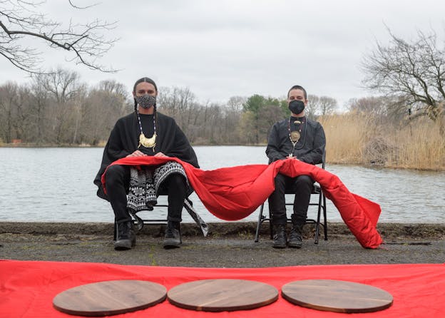 devynn emory and Joseph Pierce sit on black folding chairs on the shores of a lake, their hands on their laps looking at the camera. A red cloth is draped across both of their laps. Both wear black face masks and black clothing with long necklaces. A red cloth lies on the ground in front of them with three wooden circles on top of it.