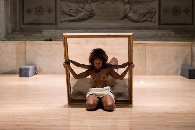A performance still of nia love supporting a wooden framed image with her back and arms. Her arms are held out in a w shape, and she looks downwards. She wears a white cloth draped across her waist and is otherwise nude. The image she holds is partially obscured but one leg and arm of a life-sized person sitting on a grey mat is visible. The image has the same beige marble background as the wall behind love. Behind the marbled wall, there is a grey stone wall carved with draped cloth around a block of text. The floor is made of light colored wood. 